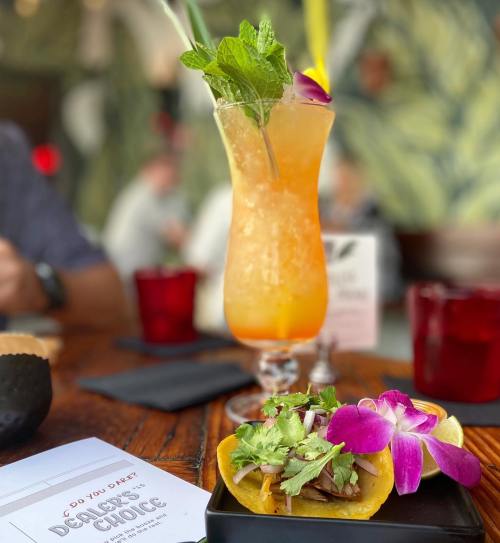A vibrant cocktail with mint and an orchid, alongside a taco topped with herbs and flowers on a wooden table.