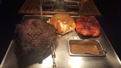 Three types of cooked meat on a serving tray: a dark, smoky piece, a golden-brown roast, and a glazed ham.