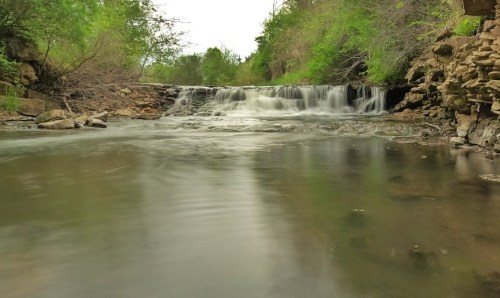 A serene waterfall cascades over rocks into a calm river, surrounded by lush greenery and trees.