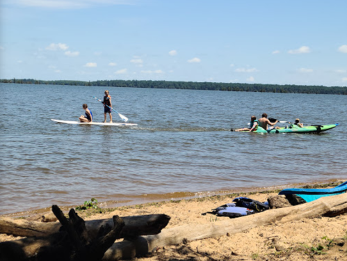 Two people paddleboarding and kayaking on a calm lake, with a sandy shore and logs in the foreground.
