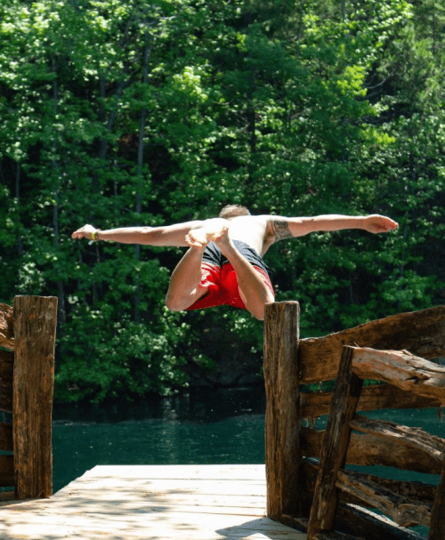 A person in red shorts leaps off a wooden dock into a serene, green forested lake.