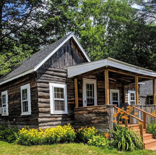 A charming log cabin with a porch, surrounded by vibrant flowers and lush greenery.