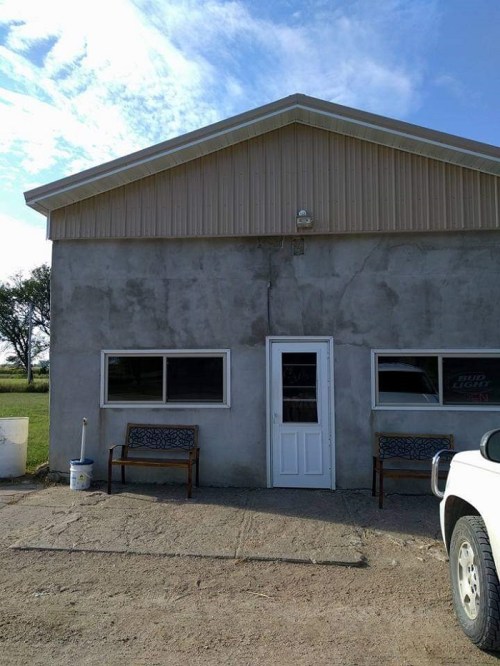 A simple concrete building with a metal roof, featuring a door and two benches outside. Clear blue sky above.