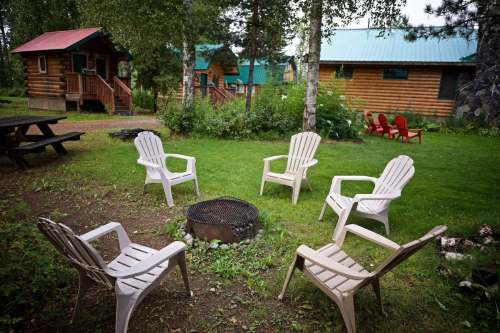 A cozy outdoor area with white chairs around a fire pit, surrounded by green grass and rustic cabins in the background.