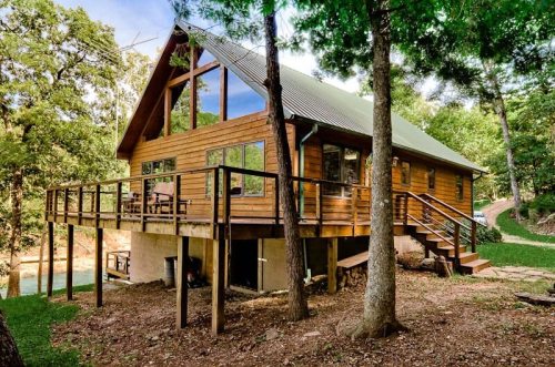 A wooden cabin with large windows, elevated on a hillside, surrounded by trees and a grassy area.