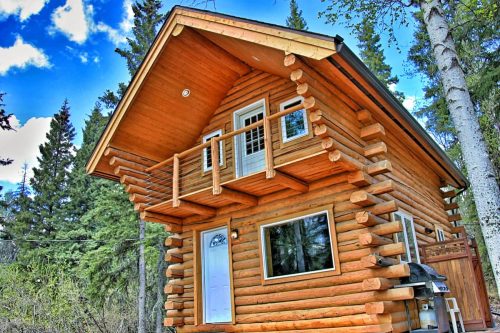 A charming log cabin with a balcony, surrounded by trees and a blue sky with clouds.