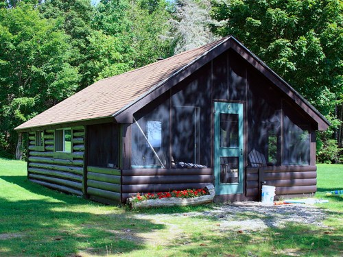 A rustic log cabin with a green door, surrounded by lush greenery and a flower bed in front.