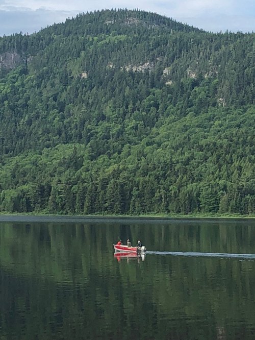 A red boat with two people glides across a calm lake, surrounded by lush green mountains and trees.