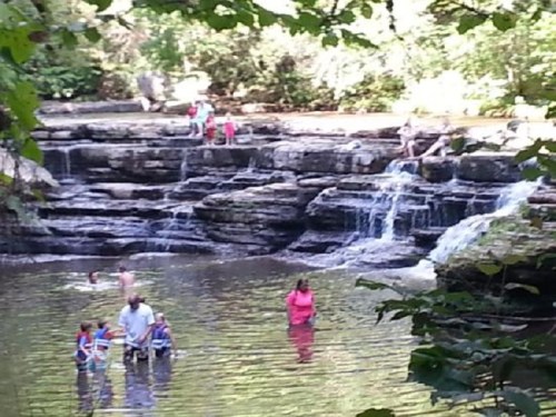 People enjoying a natural swimming area with rocks and a small waterfall surrounded by lush greenery.