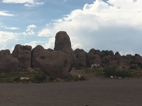 A cluster of large boulders under a cloudy sky, with a small RV parked nearby on a gravel road.