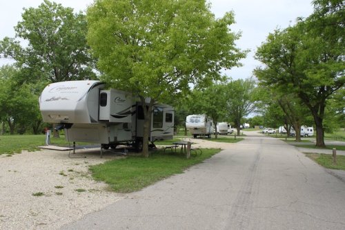 Two RVs parked under trees in a grassy campground, with a gravel path leading through the area.