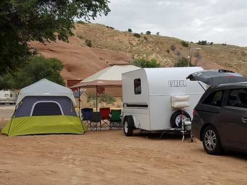 A small white camper trailer parked next to a green and gray tent, with camping chairs set up nearby in a sandy area.