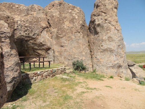 A rocky landscape featuring large boulders and a stone bench surrounded by grass under a clear blue sky.
