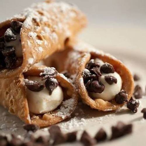 Three cannoli filled with cream and chocolate chips, dusted with powdered sugar, on a plate with chocolate chips scattered around.
