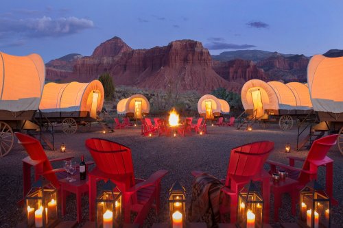 Cozy camp setup with illuminated wagons, red chairs, and a fire pit against a scenic mountain backdrop at dusk.