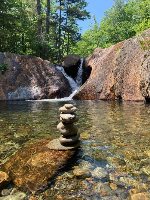A serene scene of a small waterfall surrounded by rocks and trees, with a stack of stones balanced in the foreground.