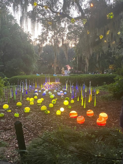 Colorful glass sculptures illuminated in a garden at dusk, surrounded by greenery and soft lighting.