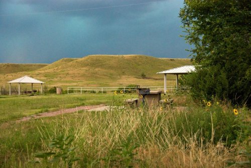 A grassy area with two gazebos, a picnic table, and sunflowers, set against a backdrop of rolling hills and darkening skies.