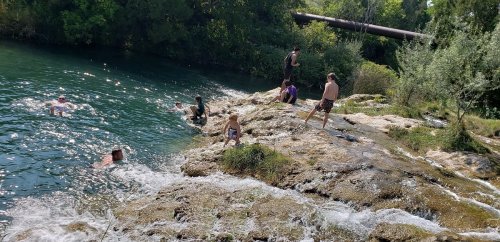 People swimming and playing by a rocky riverbank surrounded by greenery on a sunny day.