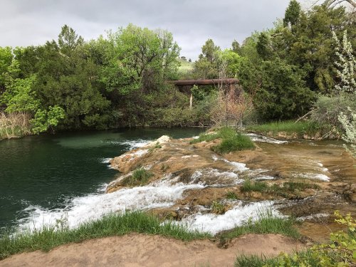 A serene river scene with flowing water over rocks, surrounded by lush greenery and a cloudy sky.