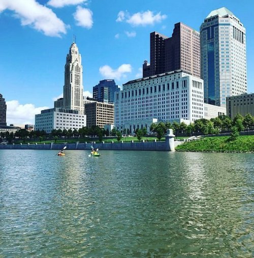 Two kayakers paddle on a river with a city skyline featuring tall buildings and a clear blue sky in the background.
