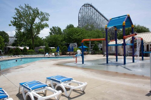 A pool area with lounge chairs, a water play structure, and a roller coaster in the background on a sunny day.