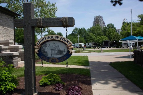 Sign for Lighthouse Point with greenery and amusement park rides in the background on a sunny day.