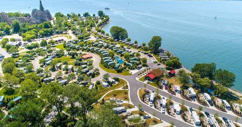 Aerial view of a lakeside RV park with green trees, winding roads, and parked RVs along the water's edge.