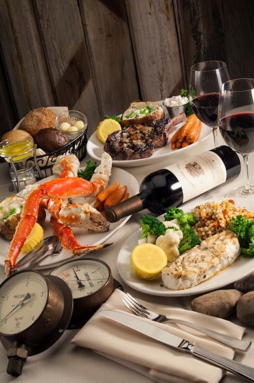 A beautifully arranged table with seafood, steak, vegetables, wine, and decorative elements, set against a rustic backdrop.
