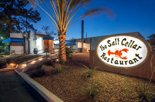 A well-lit sign for The Salt Cellar Restaurant at dusk, surrounded by palm trees and desert landscaping.