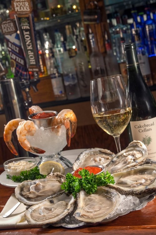 A seafood platter with oysters, shrimp cocktail, a glass of white wine, and a bottle on a bar counter.