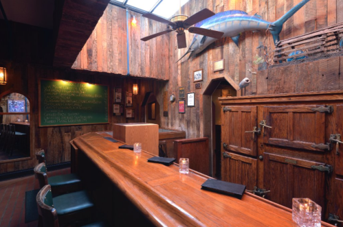 Cozy wooden bar with a ceiling fan, a marlin mounted on the wall, and a chalkboard menu in a rustic setting.