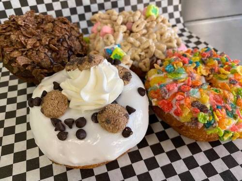 Four colorful donuts topped with various cereals, chocolate chips, and whipped cream on a checkered background.