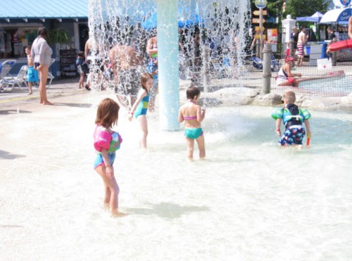 Children playing in shallow water near a fountain at a water park on a sunny day.