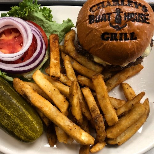 A plate with a grilled burger, lettuce, tomato, onion, fries, and a pickle, featuring the restaurant's logo on the bun.