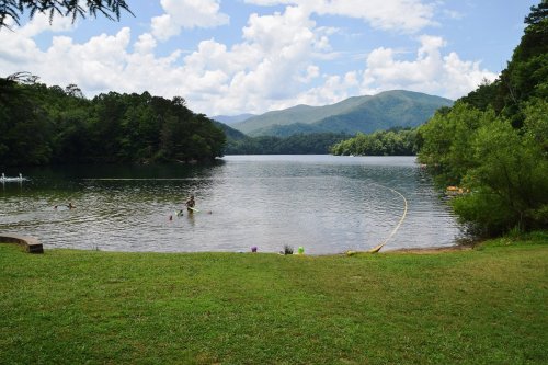 A serene lake surrounded by mountains, with people swimming and a grassy shore under a partly cloudy sky.