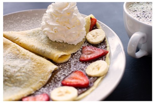 A plate of crepes filled with chocolate, topped with whipped cream, strawberries, and bananas, alongside a cup of coffee.