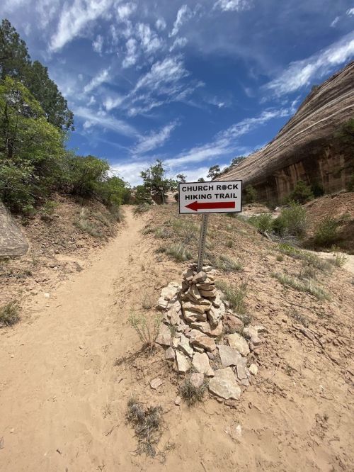 A sandy hiking trail with a sign pointing to "Church Rock Hiking Trail" amidst rocky terrain and greenery.