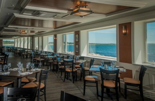 Bright restaurant interior with wooden tables and chairs, large windows overlooking a scenic waterfront view.