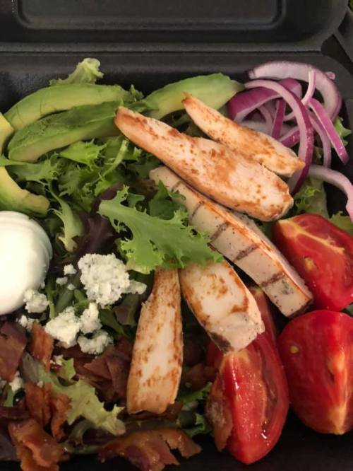 A salad with grilled chicken, avocado, tomatoes, red onion, bacon, blue cheese, and mixed greens in a takeout container.