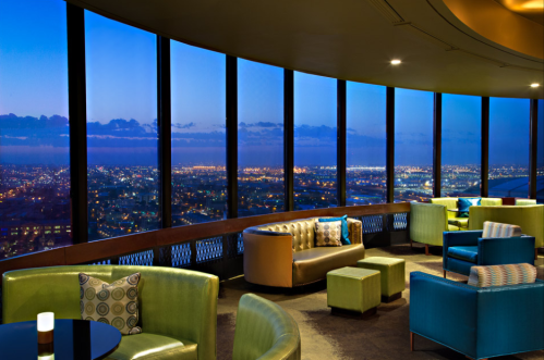 A stylish lounge with green and blue seating, offering a panoramic view of a city skyline at dusk.