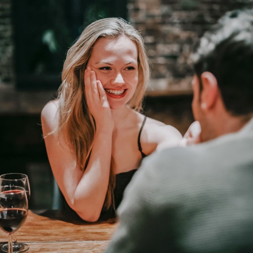 A woman smiles warmly at a man across the table, with wine glasses in the background, creating a cozy atmosphere.