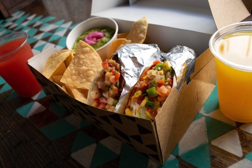 A takeout box with two tacos, chips, guacamole, and drinks on a colorful table.