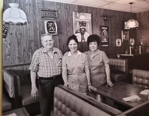 A black-and-white photo of three people smiling in a cozy restaurant with wooden walls and vintage decor.