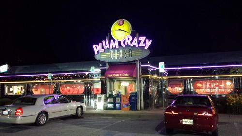 A brightly lit diner at night with a neon sign reading "Plum Crazy Diner" and parked cars in front.