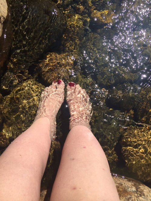Feet with red nail polish submerged in clear water over smooth stones, reflecting sunlight.