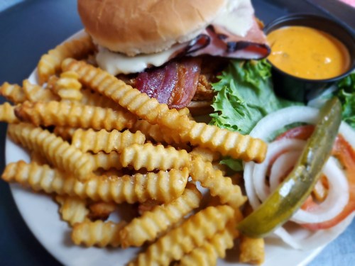 A plate with a crispy chicken sandwich topped with bacon, crinkle-cut fries, lettuce, onion, and a pickle.