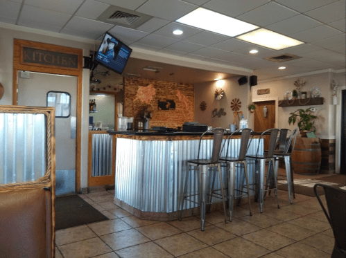 A cozy restaurant interior featuring a bar with metal stools, wooden accents, and a kitchen entrance in the background.