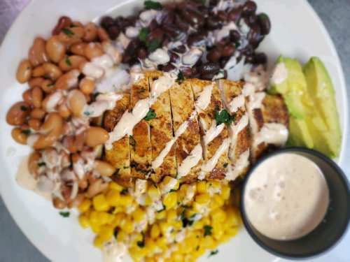 A plate with grilled fish, black and pinto beans, corn, avocado, and a creamy sauce on the side.