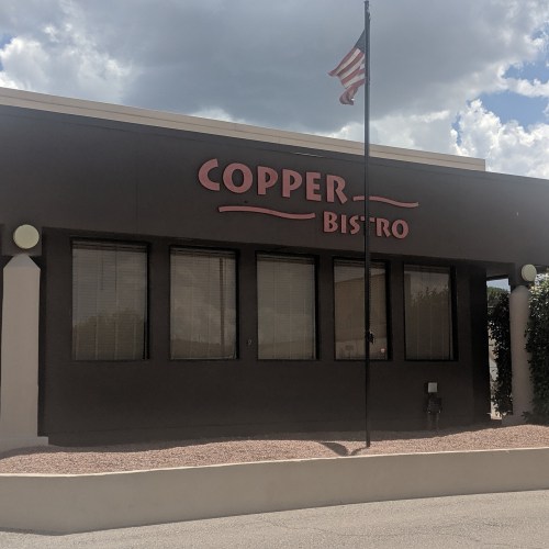 Exterior of Copper Bistro with large sign, windows, and an American flag in front under a cloudy sky.